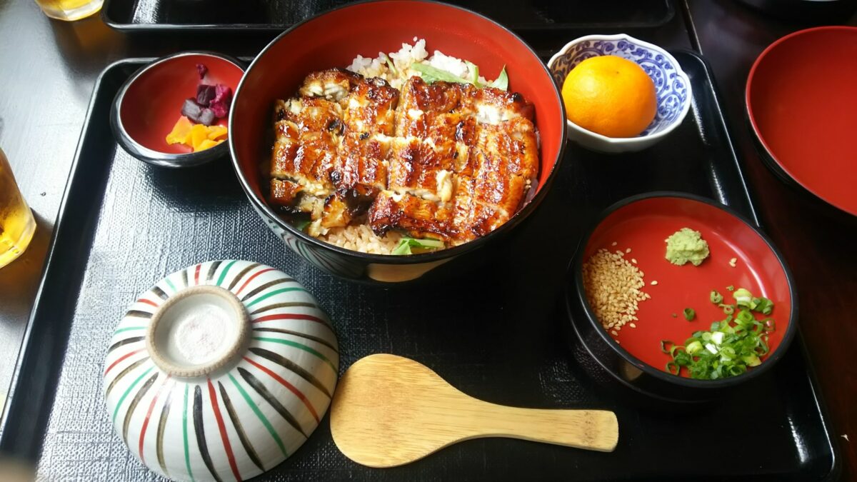 A tray of Nagoya cuisine featuring grilled eel on rice, a small bowl of wasabi and green onion, pickles, a bowl with a lid, and a wooden spoon.