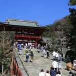 A group of people ascends a large stone staircase leading to Tsurugaoka Hachimangu Temple, a traditional Japanese building with a red façade and grey roof, set against a clear blue sky, surrounded by greenery. Nearby, the historic city of Kamakura offers charming cafes and scenic beaches.