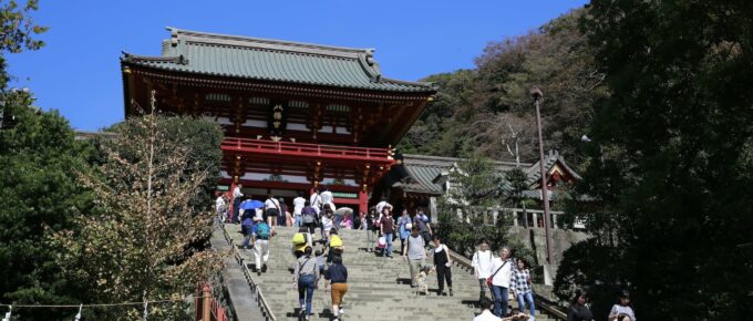 A group of people ascends a large stone staircase leading to Tsurugaoka Hachimangu Temple, a traditional Japanese building with a red façade and grey roof, set against a clear blue sky, surrounded by greenery. Nearby, the historic city of Kamakura offers charming cafes and scenic beaches.
