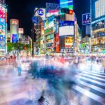 A busy Shibuya Crossing at night with colorful, bright advertisements on buildings and numerous blurred pedestrians crossing the street.