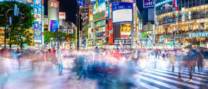 A busy Shibuya Crossing at night with colorful, bright advertisements on buildings and numerous blurred pedestrians crossing the street.
