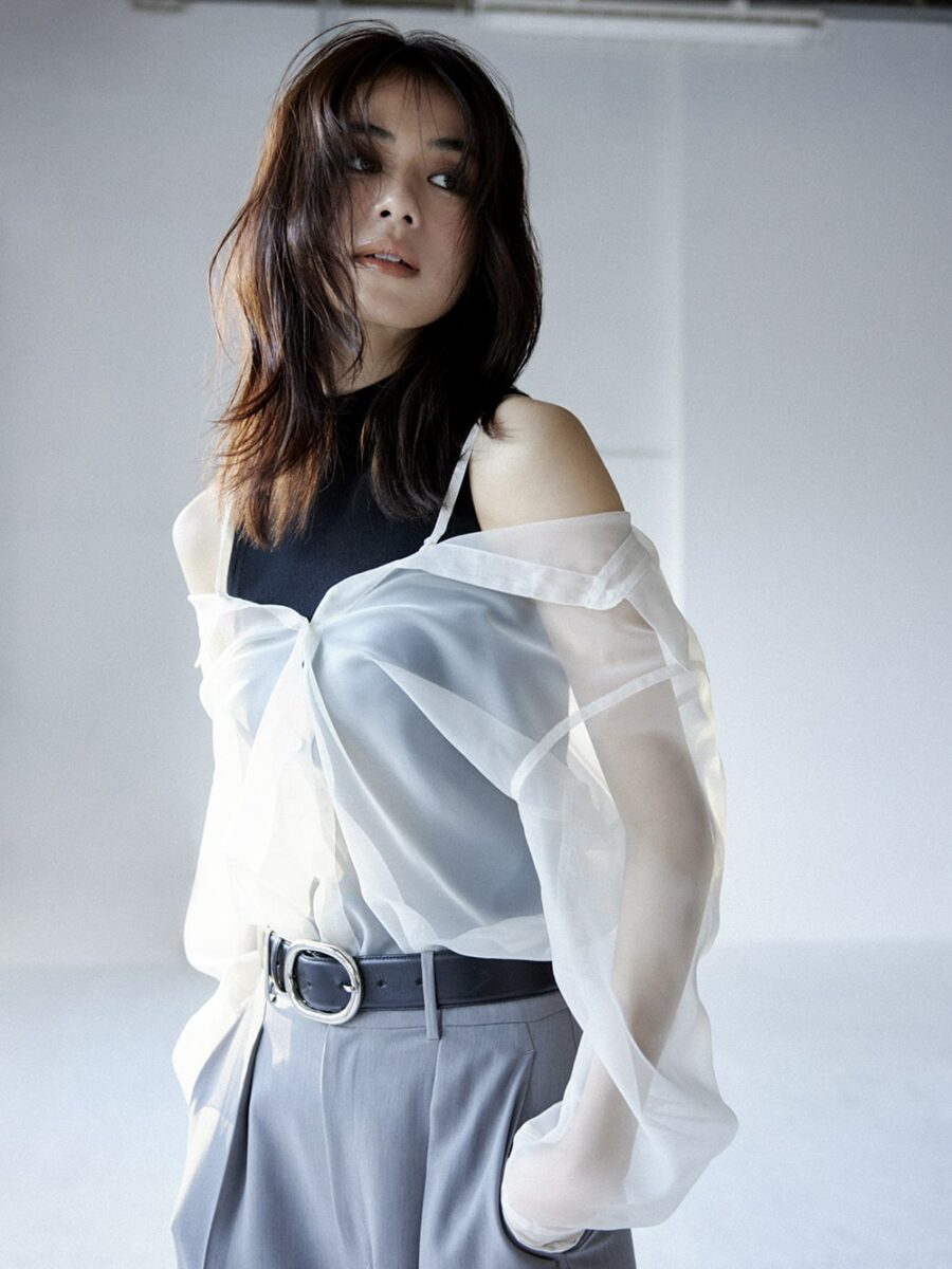 A Japanese woman wearing a sheer white blouse over a high neck black top and grey pants with a belt, sourced from popular Japanese brand Fray ID, standing against a light background.