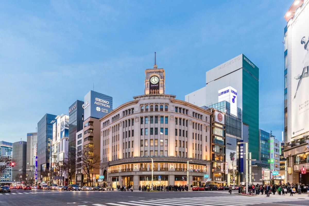 A bustling Tokyo intersection with a prominent building featuring a clock tower in the center, surrounded by various commercial buildings and billboards at dusk.