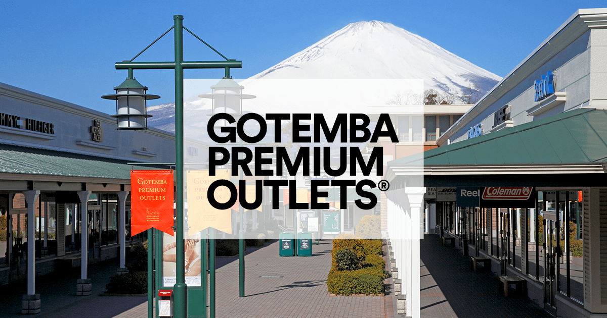 Outdoor shopping area at Gotemba Premium Outlets with various stores and streetlights, set against a backdrop of Mount Fuji on a clear day.