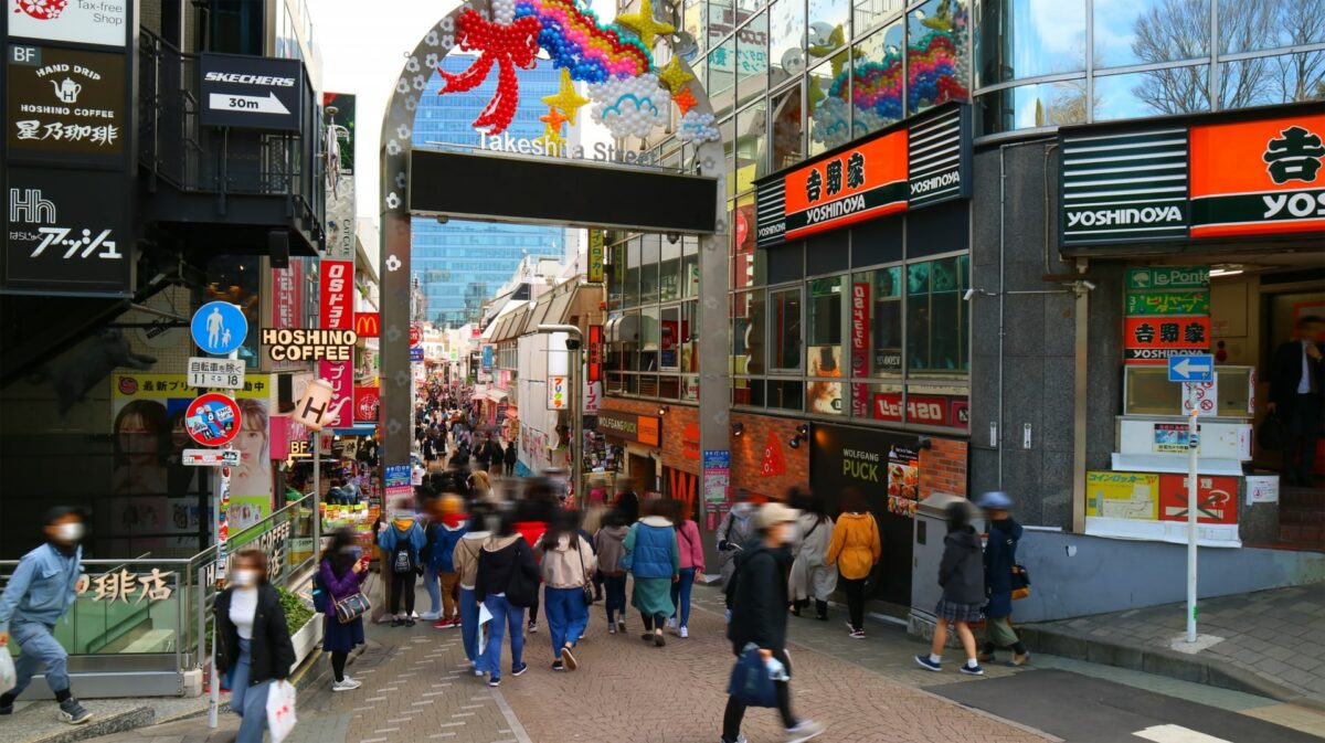 A bustling street in Harajuku, Tokyo, featuring shops with trendy clothes, colorful signs, and a crowd of people walking.