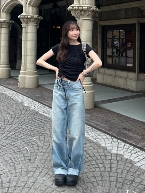 A Japanese woman stands outside a building with arches, wearing a black top, wide leg light blue jeans, and platform shoes. With long, dark hair and hands on hips.