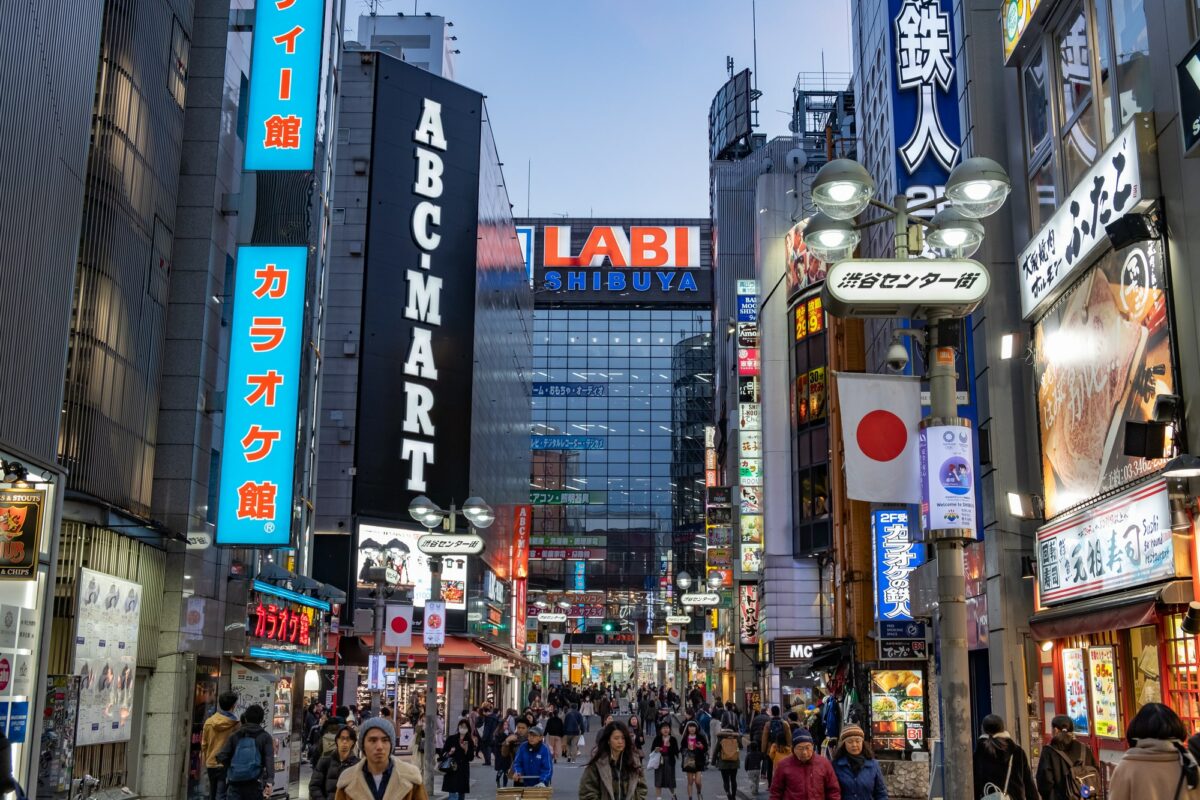 Crowded street in Shibuya, Tokyo with brightly lit storefront signs, including ABC Mart and LABI Shibuya, beckon amid towering buildings.