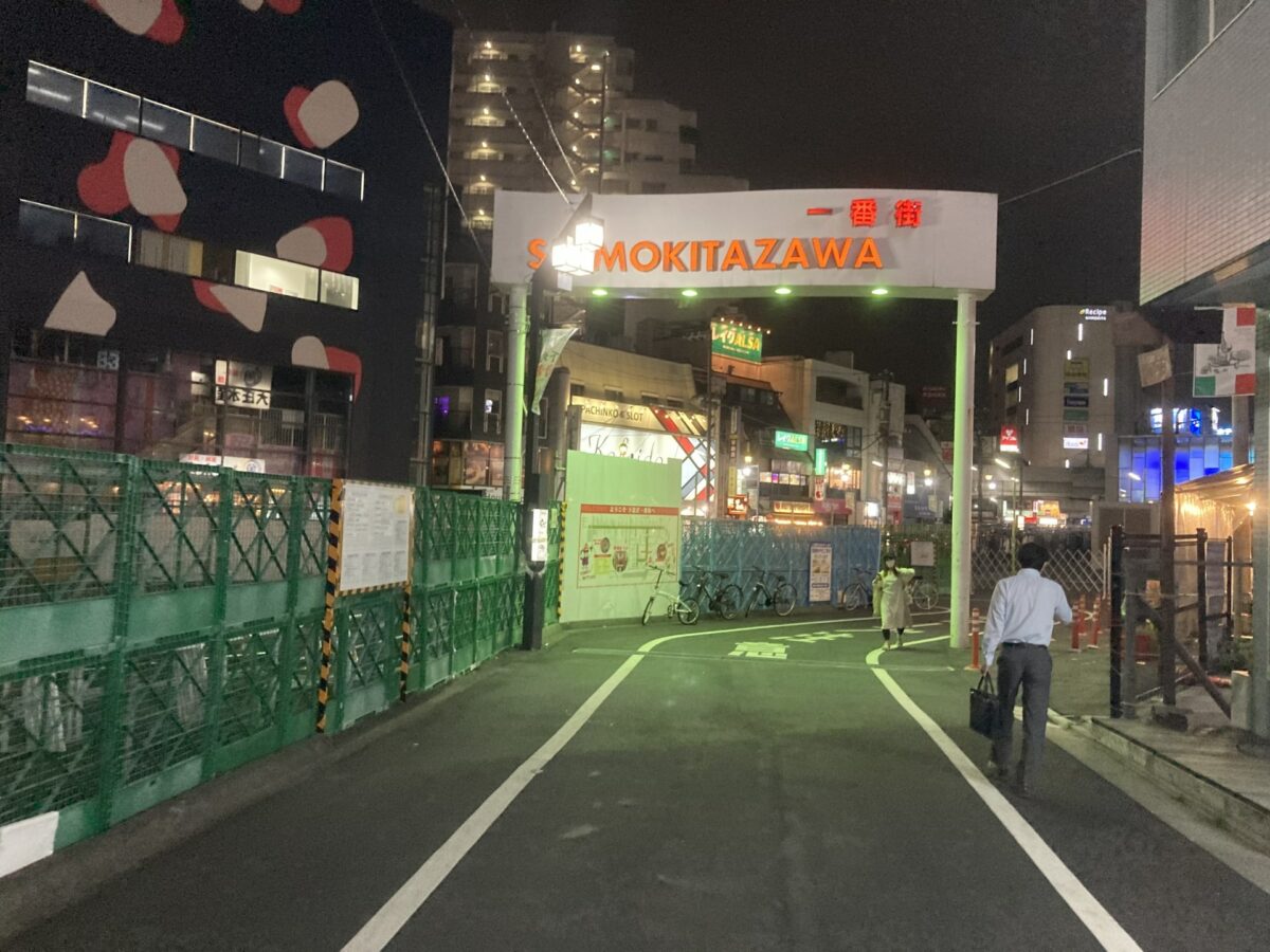 A city street at night with the sign "SHIMOKITAZAWA" lit up in the center background. A few people are walking, and buildings with illuminated windows surround the area, showcasing vintage Japanese thrift stores.