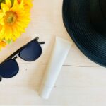 A pair of black sunglasses, a tube of Japanese sunscreen, and a black sun hat are placed on a light wooden surface next to a bunch of sunflowers.