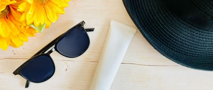 A pair of black sunglasses, a tube of Japanese sunscreen, and a black sun hat are placed on a light wooden surface next to a bunch of sunflowers.