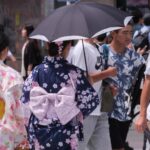 Two people in traditional Japanese attire walk among a crowd, one holding an umbrella.