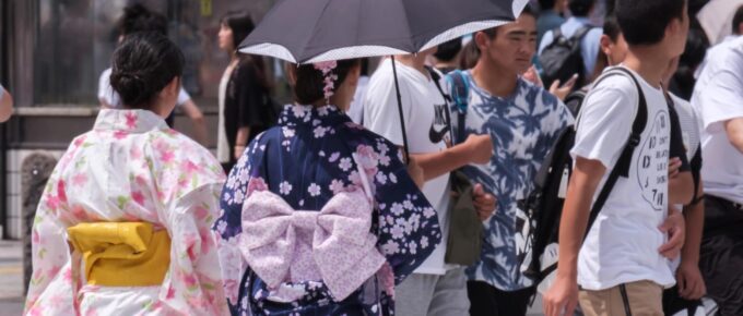 Two people in traditional Japanese attire walk among a crowd, one holding an umbrella.