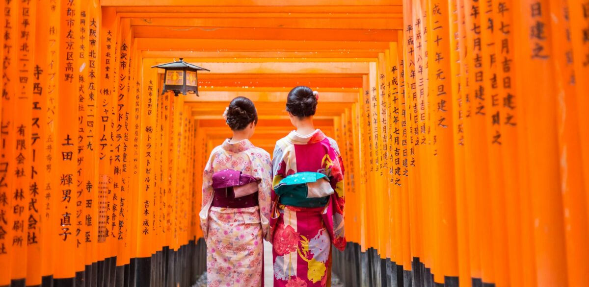 Two individuals wearing traditional Japanese kimonos walk through a corridor of vibrant orange torii gates, with Japanese characters inscribed on them.