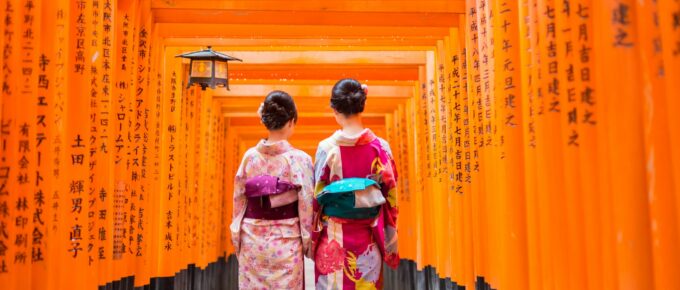 Two individuals wearing traditional Japanese kimonos walk through a corridor of vibrant orange torii gates, with Japanese characters inscribed on them.