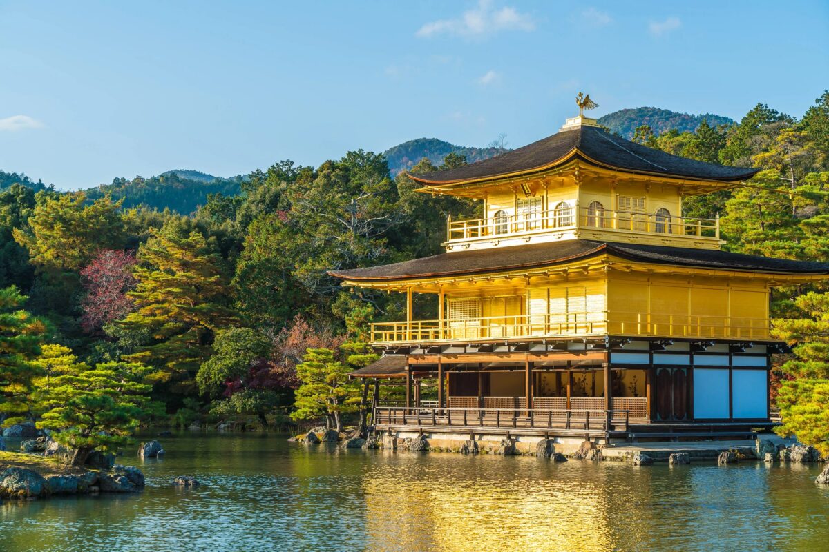 A golden temple stands by a tranquil pond, surrounded by trees with mountains in the background on a clear day.