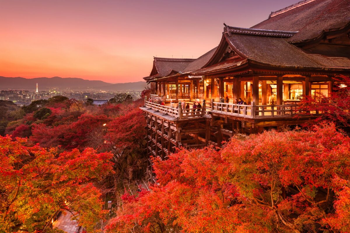 A traditional Japanese temple is surrounded by vibrant red autumn foliage at sunset with a distant Kyoto skyline in the background.