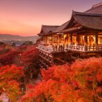 A traditional Japanese temple is surrounded by vibrant red autumn foliage at sunset with a distant Kyoto skyline in the background.