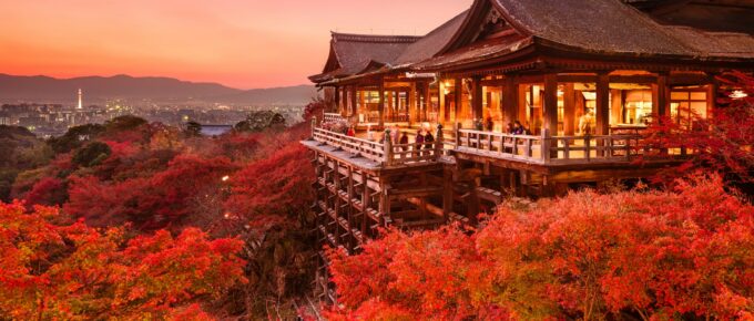 A traditional Japanese temple is surrounded by vibrant red autumn foliage at sunset with a distant Kyoto skyline in the background.