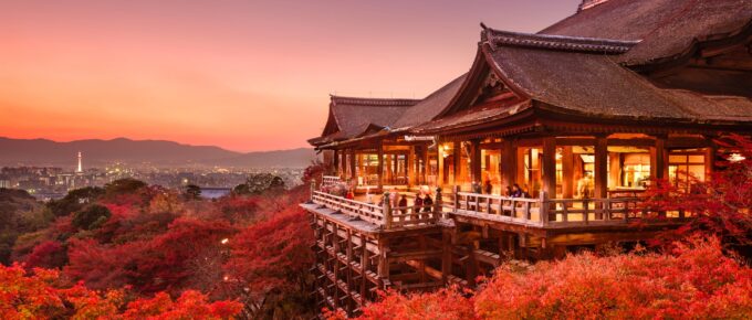 A traditional Japanese temple is surrounded by vibrant red autumn foliage at sunset with a distant Kyoto skyline in the background.