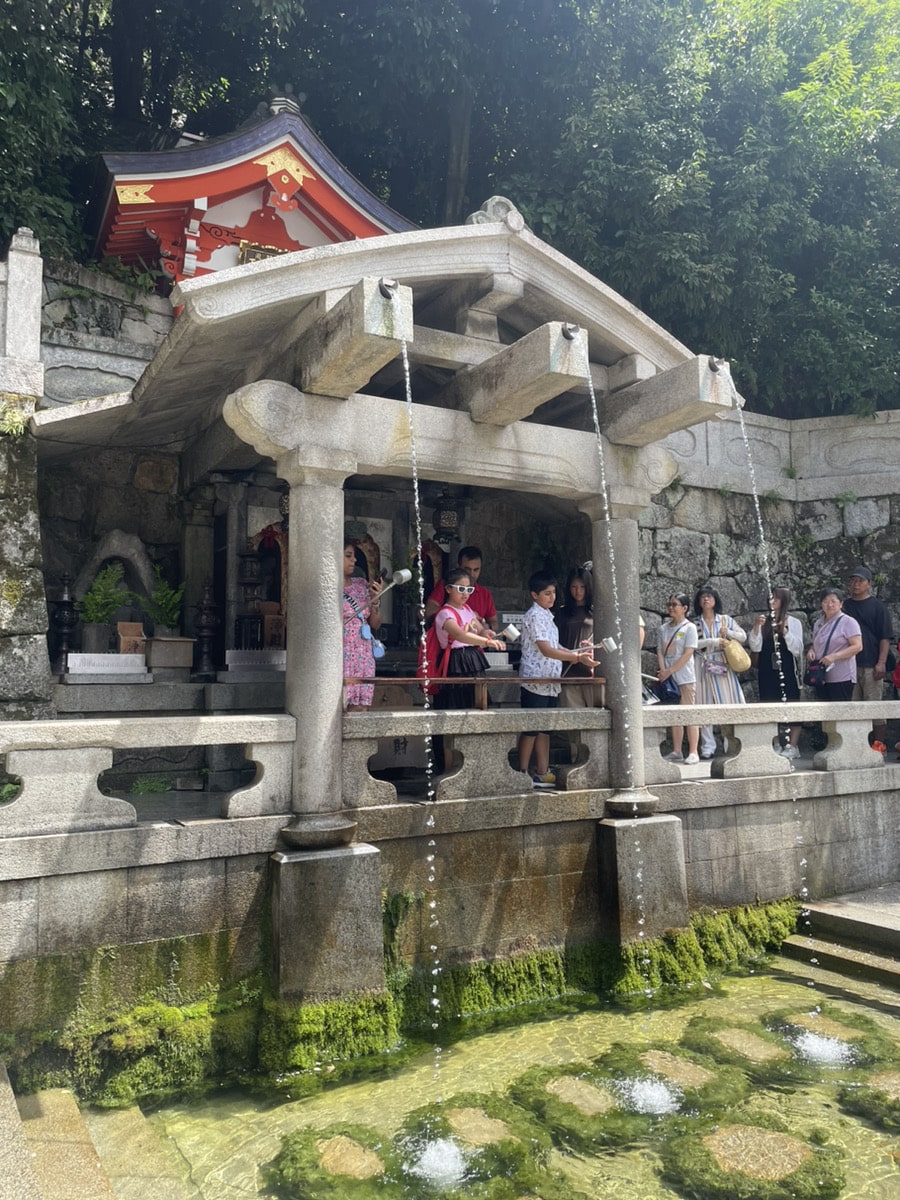 Visitors gather at a traditional Japanese pavilion with three streams of water flowing down, located in a wooded area.