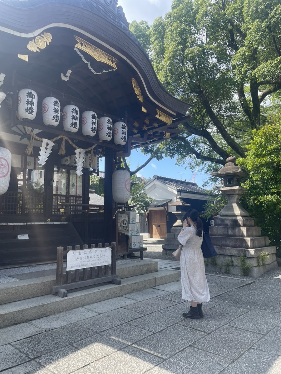 A person in a white dress stands in front of a traditional Japanese shrine, facing the structure with hands raised in prayer. Paper lanterns hang from the shrine, and trees surround the area.