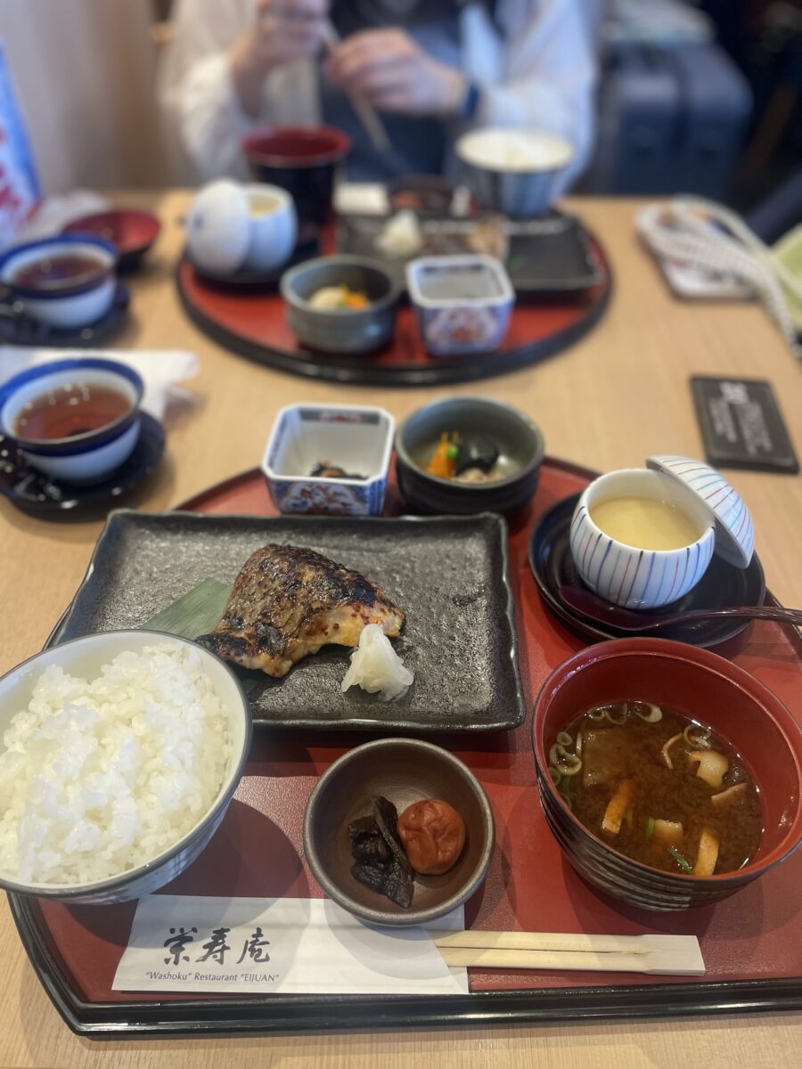 A Japanese meal set on a table in Kyoto includes grilled fish, rice, pickled plum, miso soup, and various side dishes. Two trays are visible.