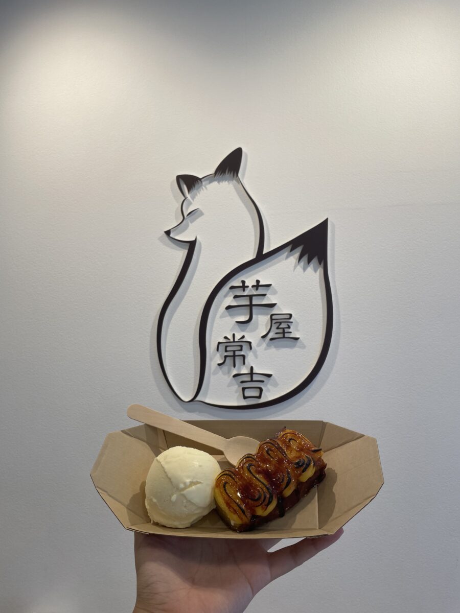 A hand holding a cardboard food tray with a scoop of vanilla ice cream and a slice of caramelized sweet potato. In the background is a wall decoration of a fox with Japanese characters.