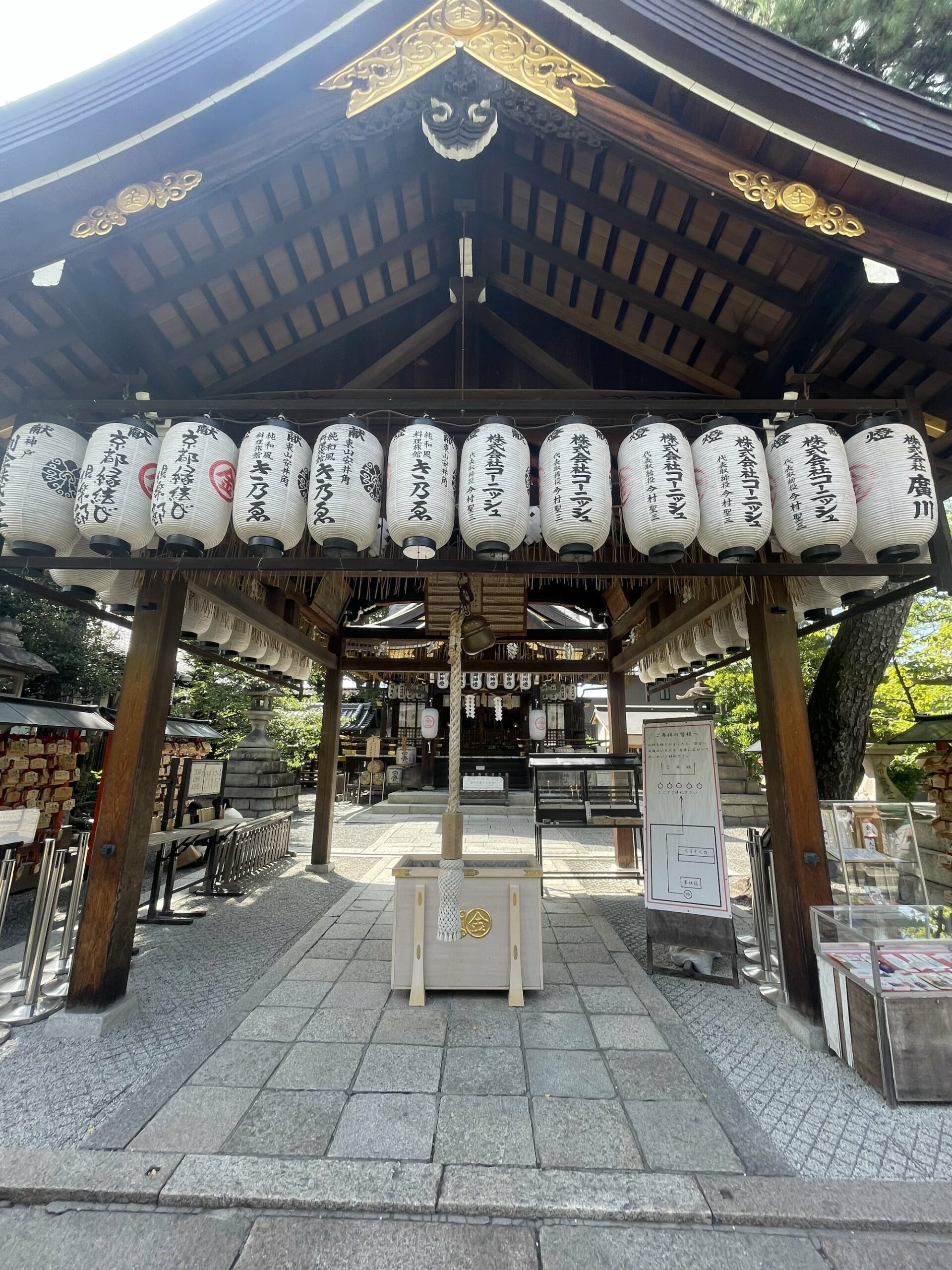 A wooden pavilion at a shrine with hanging paper lanterns, tiled roof, and informational signs. Pathway is paved with stone tiles.