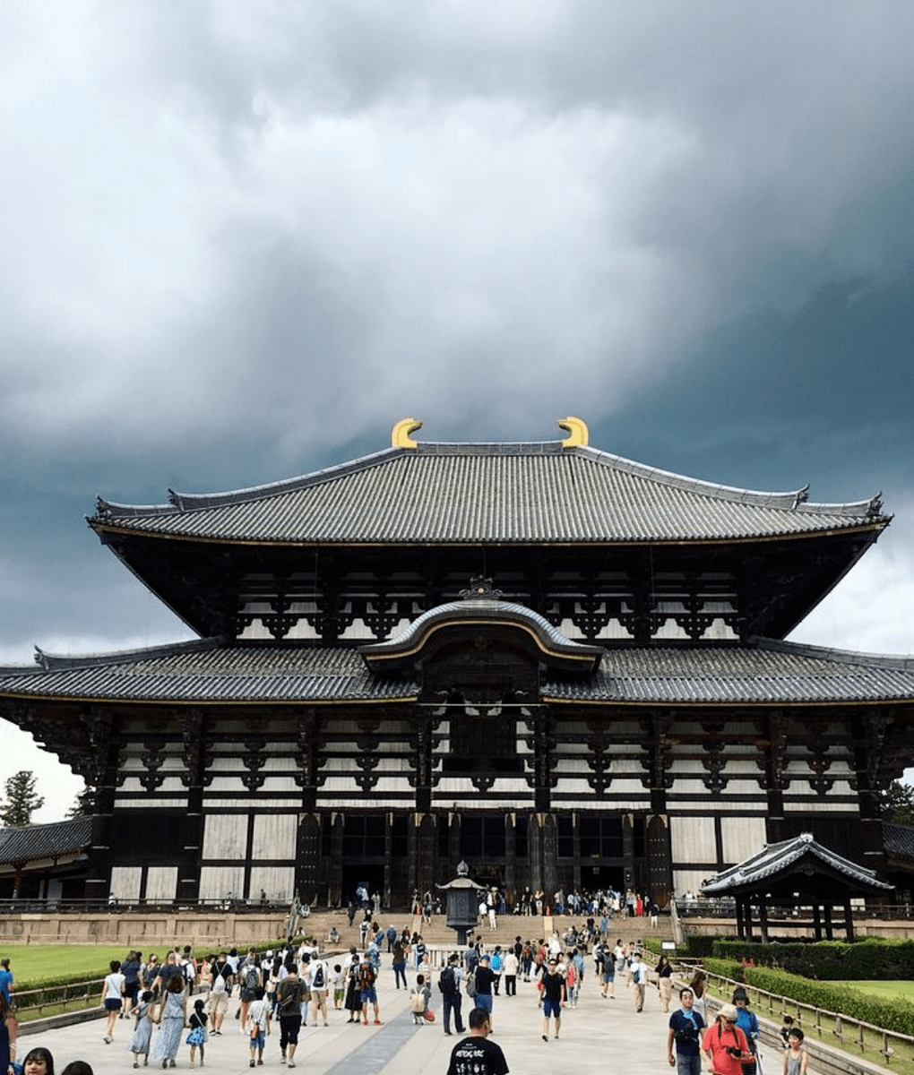 A large ancient wooden building with curved roofing stands against a cloudy sky. Many people are walking and gathering around the structure.