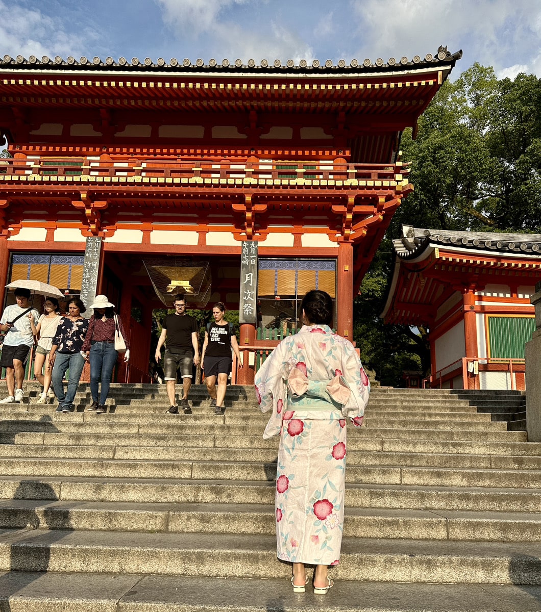 A person in a floral yukata stands at the bottom of stone steps, facing a traditional Japanese temple entrance with others descending the stairs.