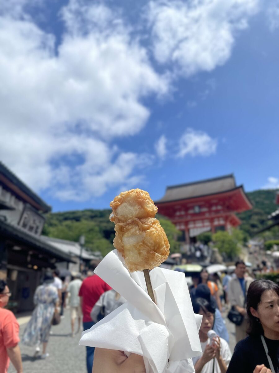 A hand holding fried food on a stick, wrapped in white paper, with a busy outdoor area in the background on a sunny day in Kyoto.