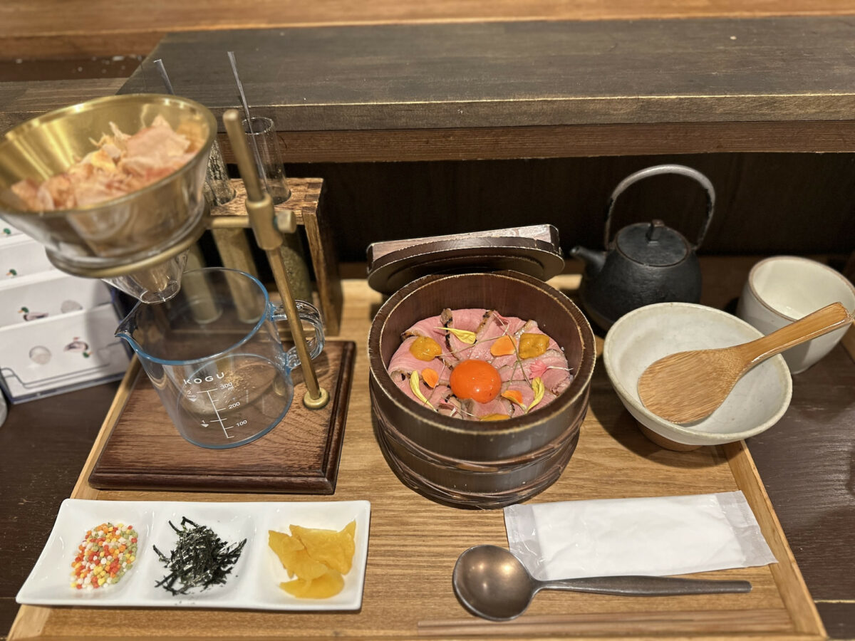 A Japanese meal including a wooden bowl with duck meat and an egg yolk, various condiments, a beaker setup, and tableware on a wooden tray.