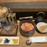 A Japanese meal including a wooden bowl with duck meat and an egg yolk, various condiments, a beaker setup, and tableware on a wooden tray.