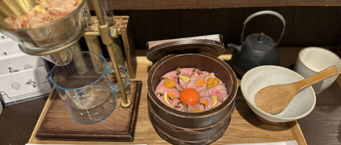 A Japanese meal including a wooden bowl with duck meat and an egg yolk, various condiments, a beaker setup, and tableware on a wooden tray.