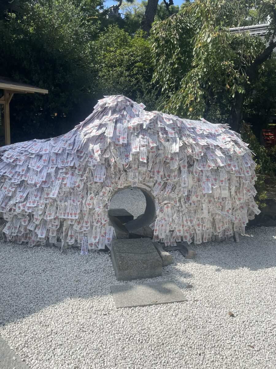 A structure covered with numerous folded papers stands in a gravel area, surrounded by greenery, reminiscent of the shrines and temples of Kyoto. A circular archway is at the base of the structure.