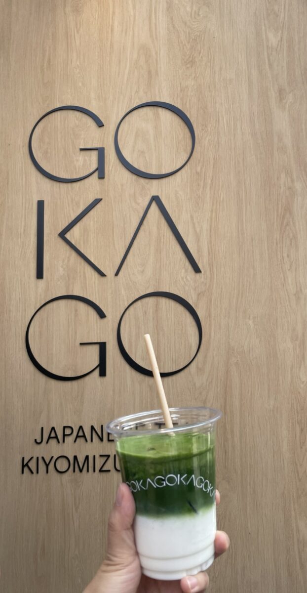 A hand holds a green and white layered drink in front of a wooden wall with the text "GOKAGO JAPAN KIYOMIZU," capturing the essence of Kyoto's must-eat foods.