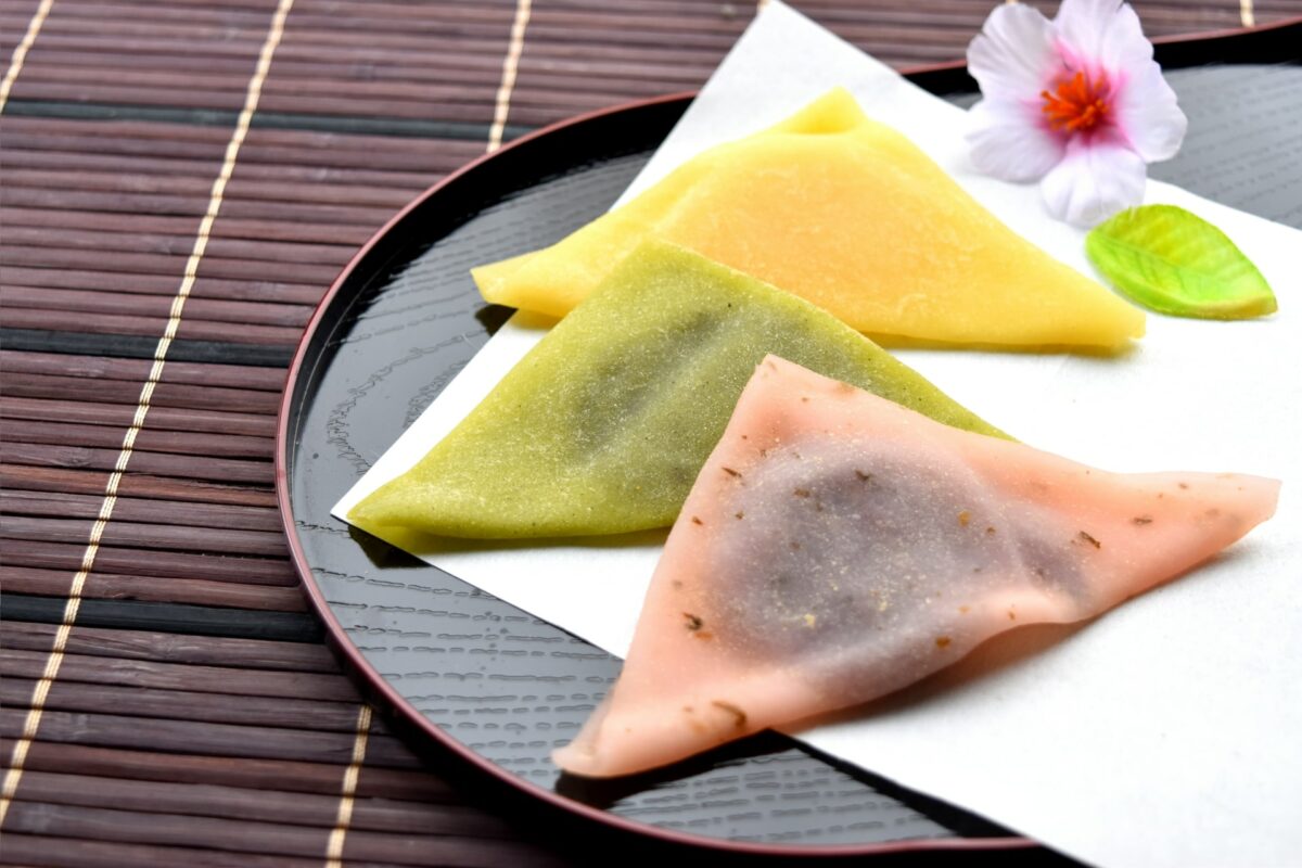 Three triangular mochi pastries in yellow, green, and pink are placed on a black tray with a white napkin. A decorative flower and leaf are next to them. The background has a bamboo mat pattern, evoking the traditional elegance of Kyoto. This delightful treat is one of Japan's must-eat foods.