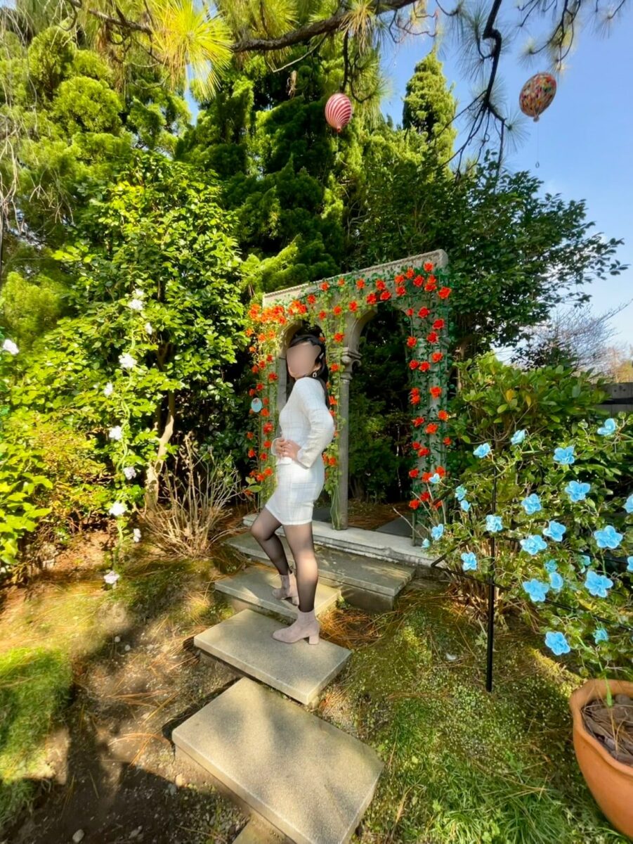 Person in a white dress and tights poses on stone steps in a garden with an arch covered in red flowers and greenery.