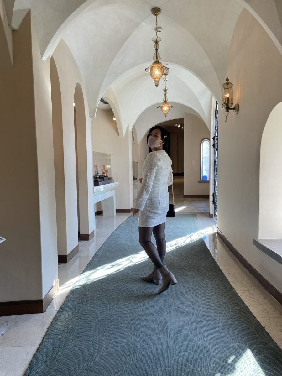 Person in a white outfit and mask stands in a hallway with arched ceilings and chandeliers, looking back.