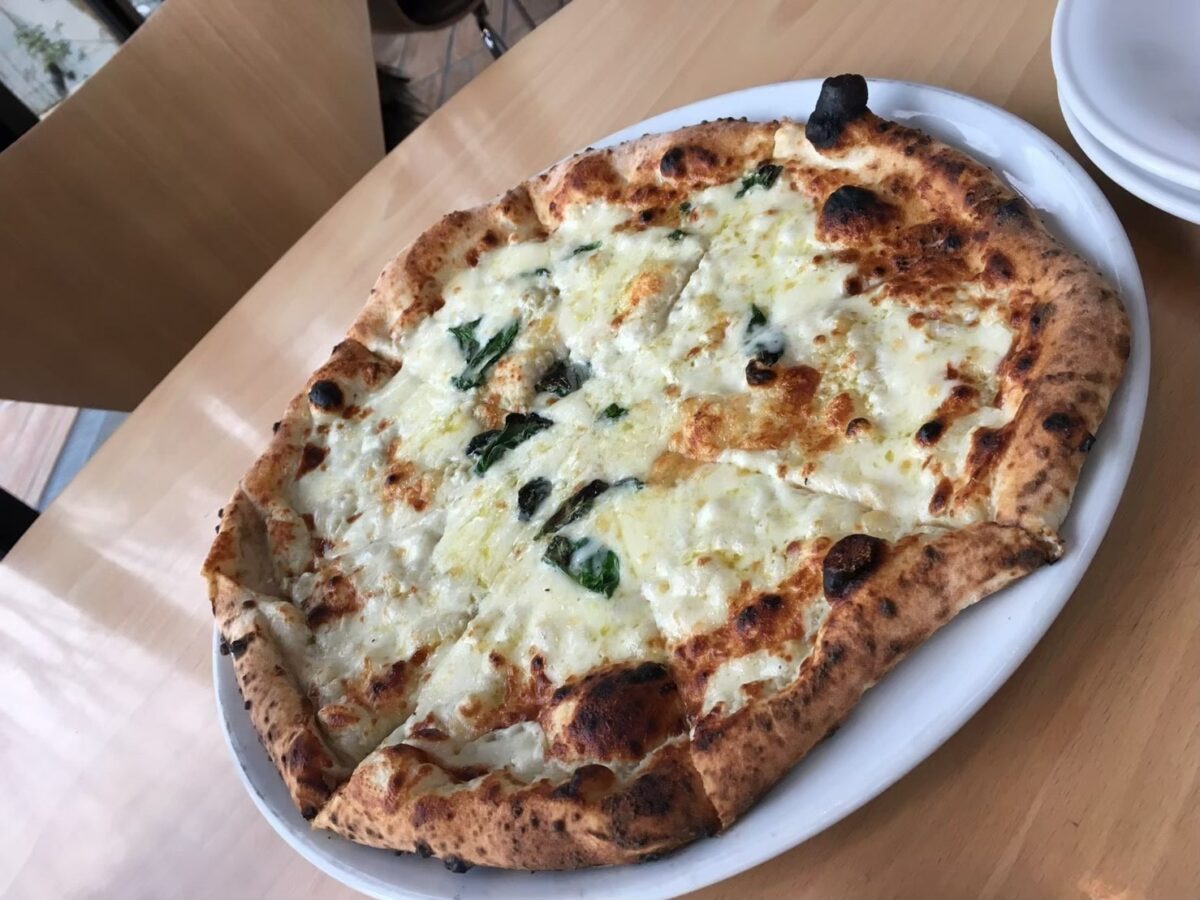 A wood-fired pizza topped with basil and melted cheese on a white plate, served on a wooden table.