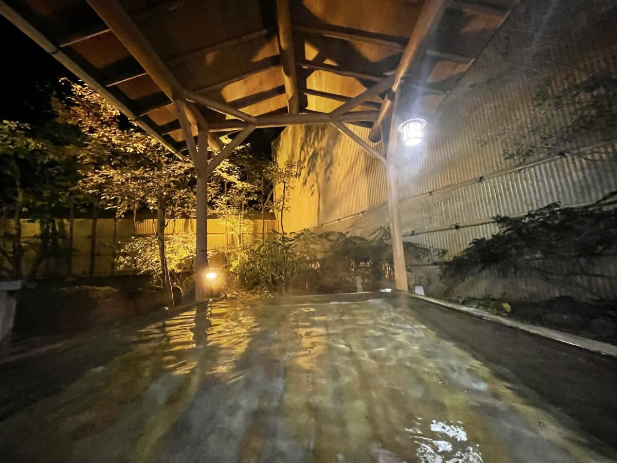 Outdoor hot spring bath at night, surrounded by a wooden structure and dimly lit by lanterns. Trees and bamboo fence in the background.