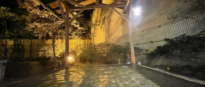 Outdoor hot spring bath at night, surrounded by a wooden structure and dimly lit by lanterns. Trees and bamboo fence in the background.