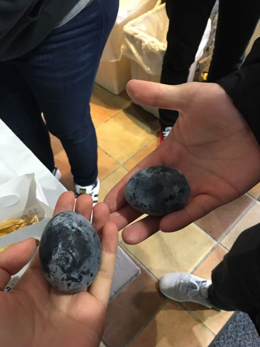 Two hands holding black eggs in a tiled room, with people standing nearby.