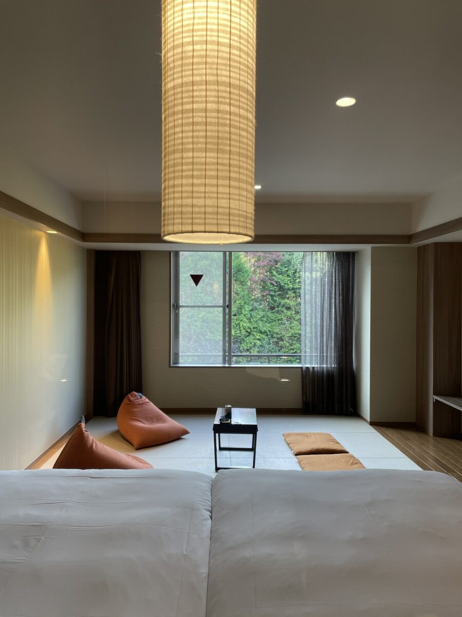 A minimalist room with a bed in the foreground, bean bags, a low table, and a window offering a view of greenery. A hanging lamp provides soft lighting.