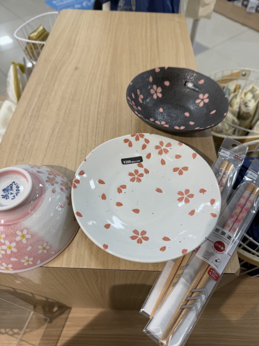 Ceramic dishes with floral patterns reminiscent of cherry blossom goods sit alongside a pack of chopsticks on a wooden display table.