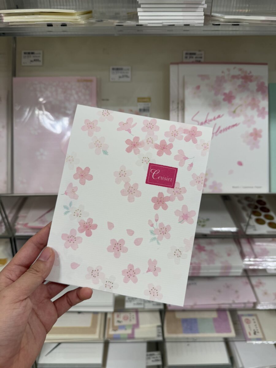 A hand holds a floral-themed notebook with pink cherry blossoms, displayed in front of shelves stocked with similar stationery items.