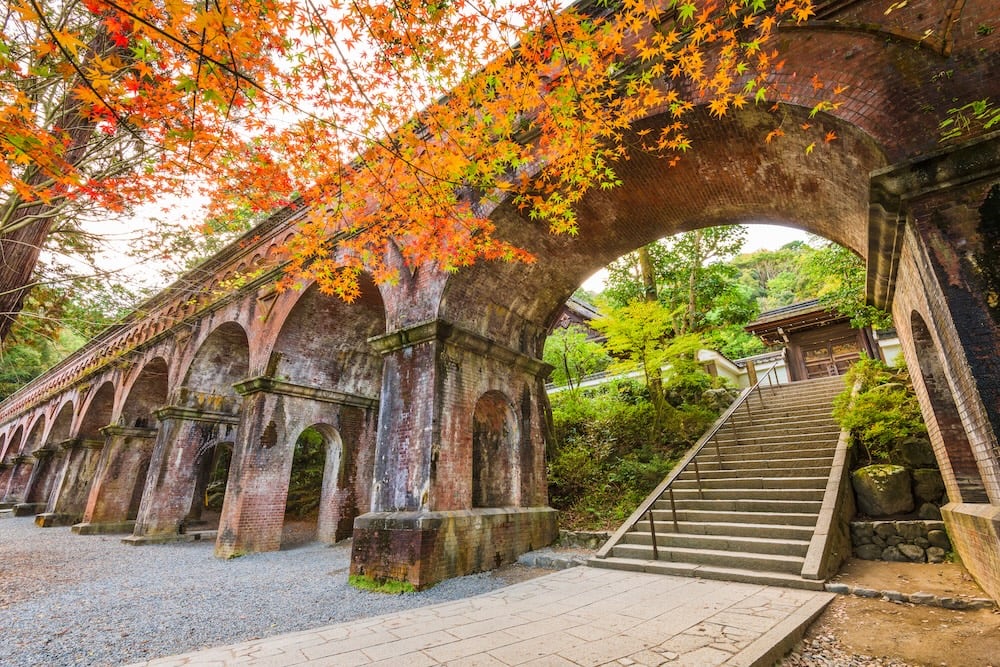 Brick aqueduct amidst autumn trees with vibrant orange leaves, a gravel path beneath, and stone stairs leading up to a wooden building.