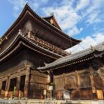 A large traditional wooden Japanese temple surrounded by trees, under a partly cloudy sky.